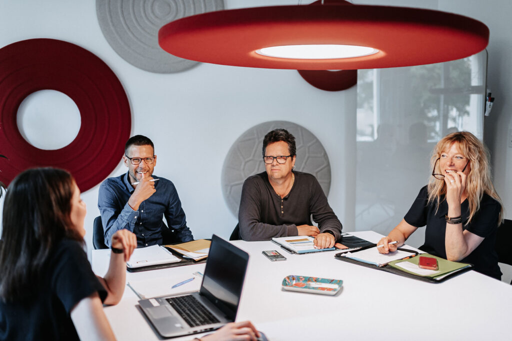 Vier Personen sitzen an einem Tisch. Sie diskutieren und machen sich Notizen auf einem Schreibblock und mittels Laptop.